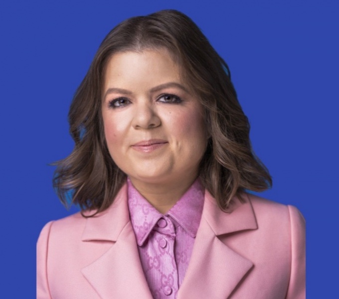 Sinead faces the camera face on. She has long dark brown hair, tucked behind her shoulders and wears a pink jacket and blouse. She stands in front of a deep blue background.