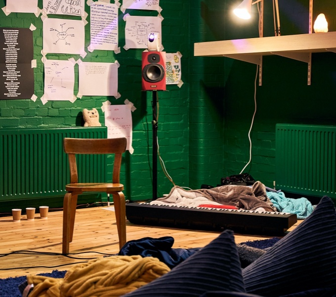 A corner of a room with green walls and a wooden floor. Central to the image is a wooden chair, to the right a large keyboard is on the floor, cushions and blankets. Behind, on the painted brick walls are large pieces of paper with writing and diagrams.