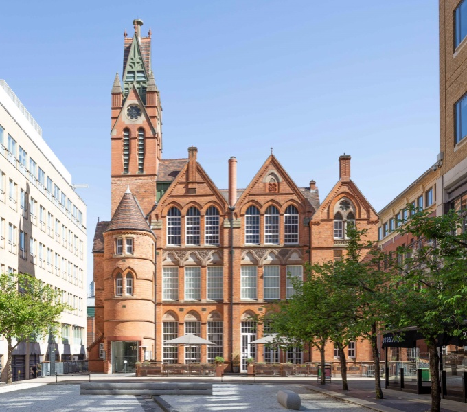 A view from the front of the Ikon gallery. A red brick building in a gothic style with tall elegant windows and clock tower on the left hand side, set against a bright blue sky. 