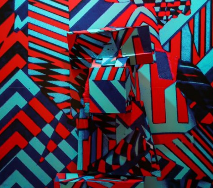 A  blue an red background of a dazzle pattern design used on warships. Artist Aaron stands in front in a 3d costume of the same design - barely visible