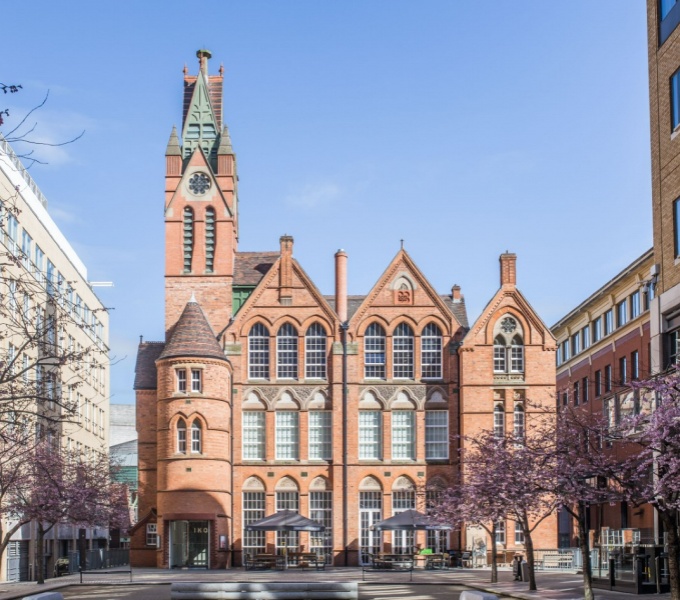 A view from the front of the Ikon gallery. A red brick building in a gothic style with tall elegant windows and clock tower on the left hand side, set against a bright blue sky. 