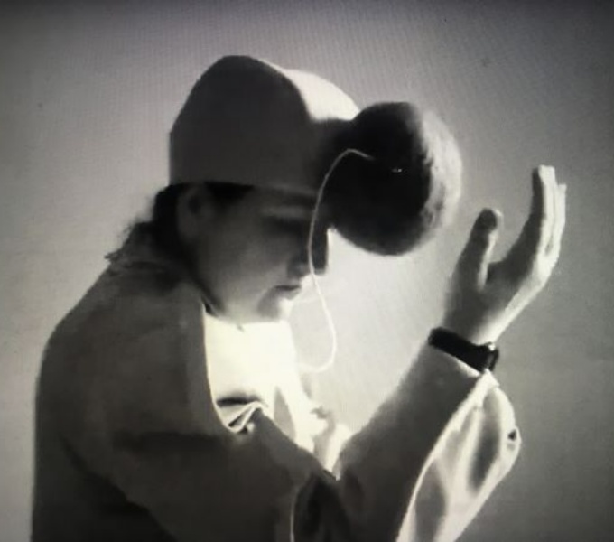 A black and white photograph of Nicola. She wears a white jacket and a light coloured cap with pom pom shaped sound equipment attached to the front. Her head tilts downwards and her right hand reaches upwards towards her head.