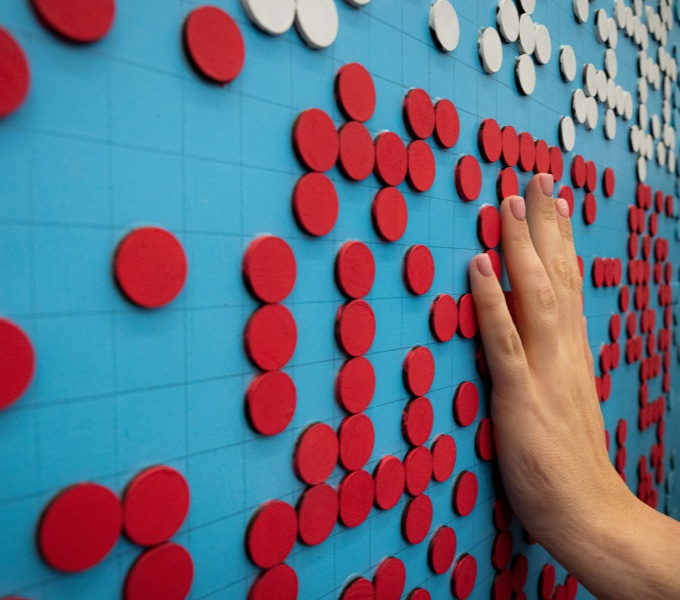 A hand is exploring a work of art made of large braille text that has been cut from painted wood. The braille is arranged in bands: a lower band of red on a bright blue background, and an upper band of white on bright blue background.