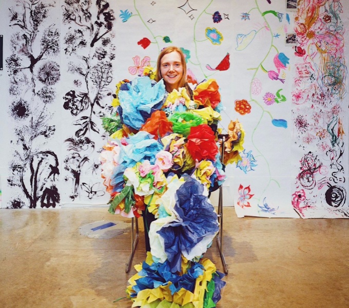 Jess Starns covered in colourful paper against the backdrop of a wall covered in both colour and black and white drawings.