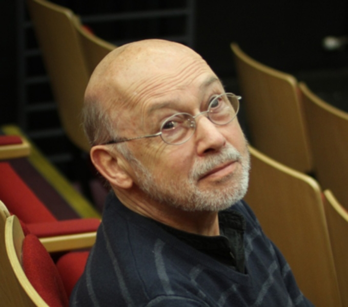 Jonathan sits on red plush theatre seat with a wooden back. He wears a dark coloured jumper and frameless glasses. He looks sideways and slightly upwards toward the camera. There is a slight twinkle in his eye.