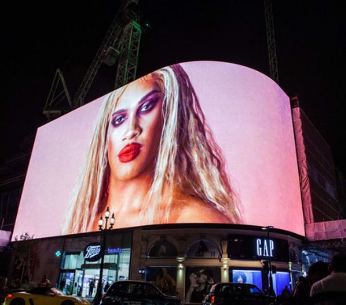 A  night scene of a bust urban street. A large screen that wraps around a building with shops below. On the screen is an image of a person with long blonde hair, bright red lipstick and bright eye make-up stood against a pink background.