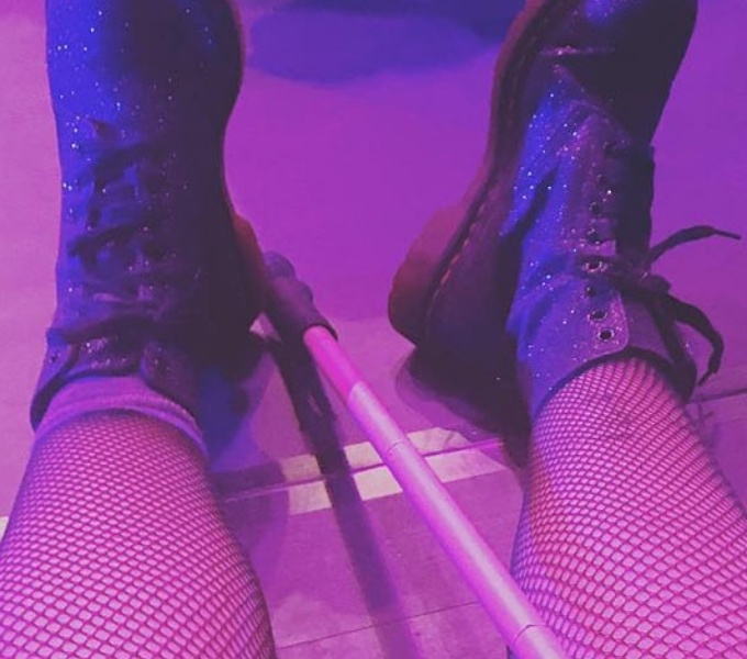 A square frame captures a medium close-up of someone sitting on the floor, Two legs adorned in delicate fishnet stockings rest gently, while sparkling rubber boots ignite a sense of daring, joy, and rebellion. Between them, a metal cane rests on their leg