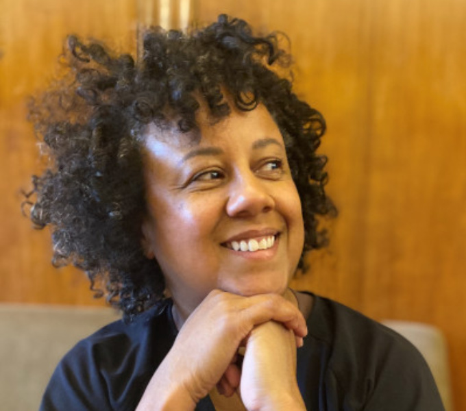 A head and shoulders photographic portrait of Lou Mensa. Her skin tone is light brown, and she has dark brown eyes. Her short, curly dark brown hair has subtle highlights and a fringe. She wears a navy blue, long-sleeved top.