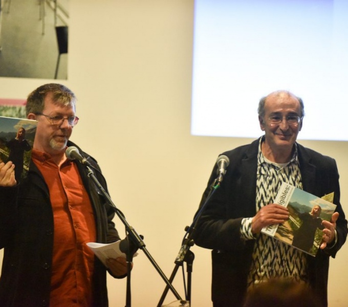 Colin stands to the left in front of a microphone wearing a brown suit and orange shirt. He holds a copy of the book The Incorrigibles up in his right hand. To his left is Mike Layward, wearing a black jacket and white and grey patterned shirt.  