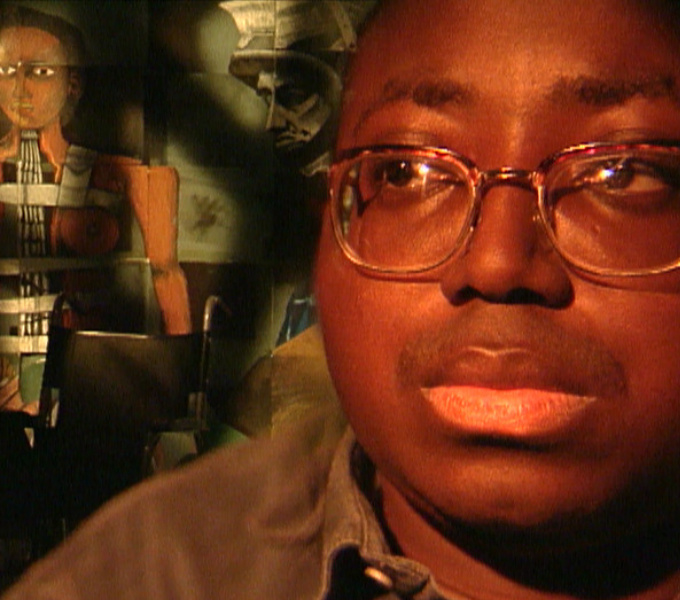 A photo of Donald Rodney. He has dark brown skin, wears gold framed glasses and a khaki shirt. An abstract painting of a figure is in the background.
