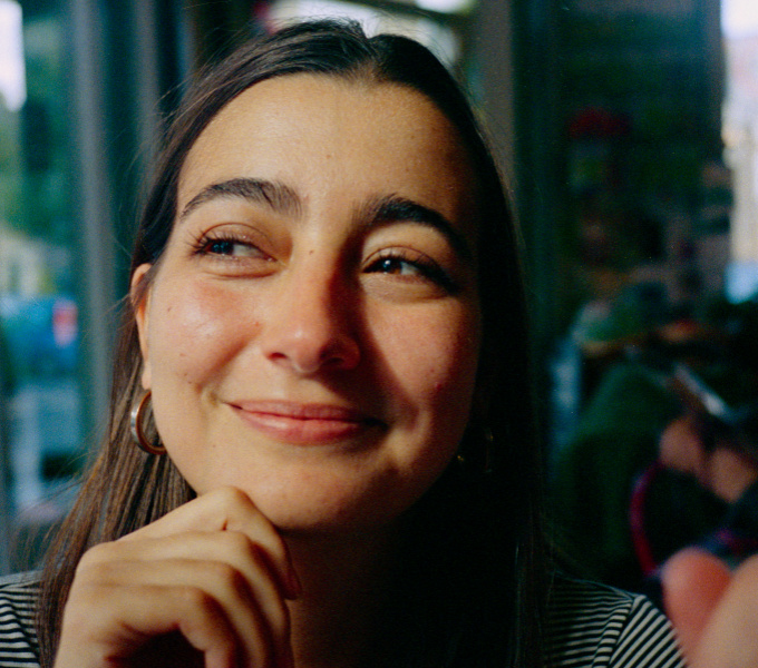 A head and shoulders, colour photographic portrait of Mariana Lemos. Mariana has shoulder length brown hair, dark brown eyebrows and brown eyes. She has tanned skin. She wears gold hooped earrings, a gold cross pendant on a chain around her neck