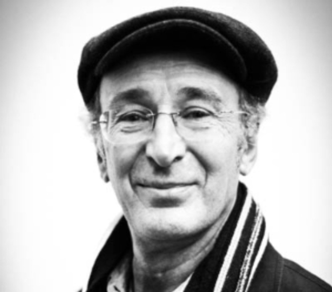 A black and white portrait photograph of Mike Layward. He wears a stripy scarf, thin framed glasses and cap. He smiles as he looks at the camera. 