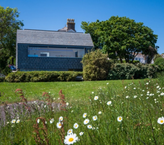 A rectangular grey stone building with slate roof and a long gallery picture window is situated amongst green trees and shrubbery. To the front is a verdant grassed lawn. There are wildflowers in the foreground. 