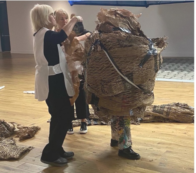 Disabled Artist Grace Currie stands in a gallery space and is wrapped in layers of packaging by two women. Grace's feet protrude from the bottom of the package. Her face, head, body and hands are all obscured. The performance was part of WAIWAV.