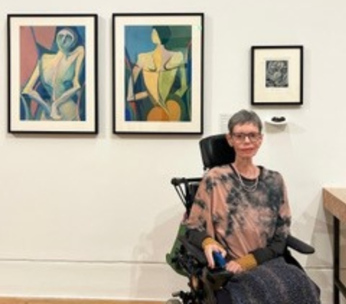 A cropped photo of artist, Nancy Willis is seated in a wheelchair in front of her paintings, on a white wall to the left. She has short cropped hair, and wears gasses and a pink and grey tie-dye top.