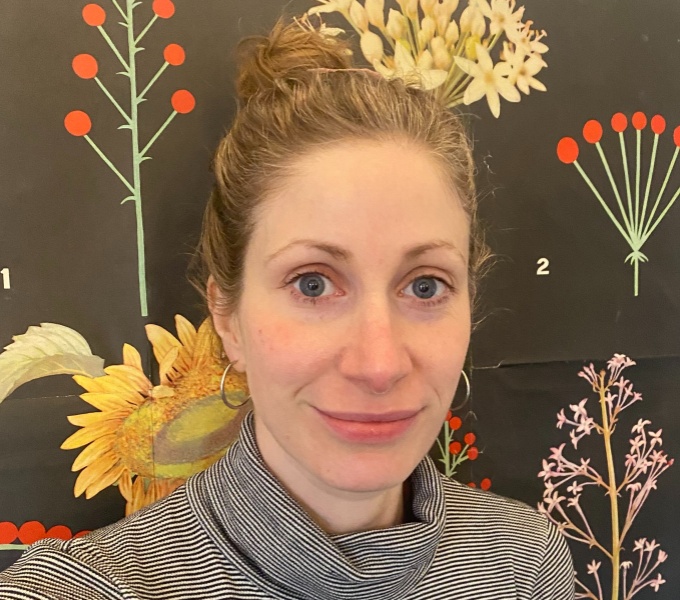 Headshot of Nathalie, a white woman with blue eyes and dark blonde hair tied up into a bun. She is looking directly at the camera and is smiling. She is wearing a striped polo neck and silver hoop earrings. 