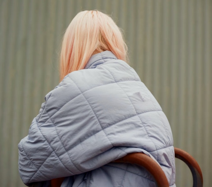 A young person with long blonde hair and a grey puffer jacket sit on a wooden cafe chair, they sit with their back to the camera.