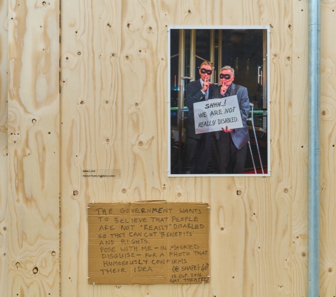 An image of two people holding a sign saying 'Shhh...! We are not really disabled', mounted on a piece of plywood.