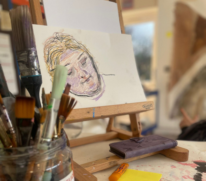 A photograph of a portrait drawing of a woman with blonde hair resting on a tabletop easel. in the foreground is a jar of pant brushes labelled 'big brushes'. There are some smaller drawings on the table too, along with a mobile phone in a brown case.