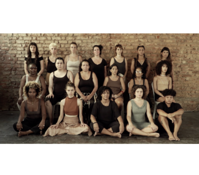 a group photograph of eighteen people (who appear to be women) arranged in three rows, the back row standing, the middle row seated on chairs and the front row seated on the floor. The subjects, all various ethnicities, are wearing muted coloured clothing