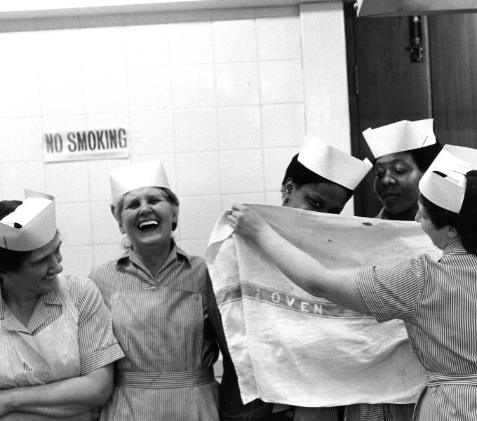 A black and white image of dinner ladies in stripey dresses and white paper hats laughing, as they joke around with tea towels. 