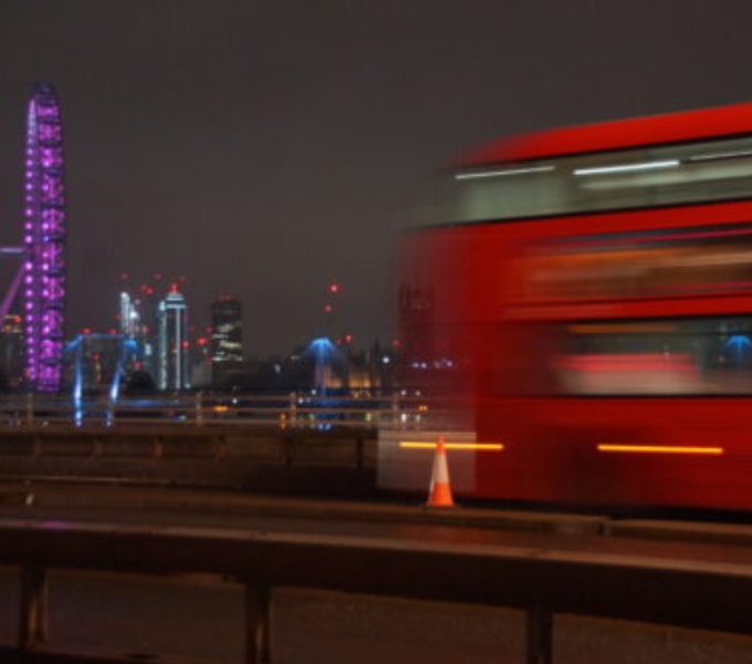 A blurred image of a red double decker bus moving across a city bridge. Its night time and the lights of tower blocks shine in the distance.
