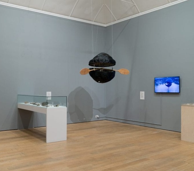 A blue grey painted gallery space with a wooden floor and white vaulted ceiling. A white table with glass cabinet is to the left, a dark object in two halves hangs from the ceiling, with oar like shapes at the centre. A screen is on the righthand side.