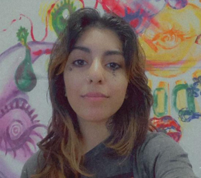 Sanaa has mid length dark brown hair and wears a dark grey t-shirt with red motif. Behind her is a white wall painted in graffiti like illustrations.
