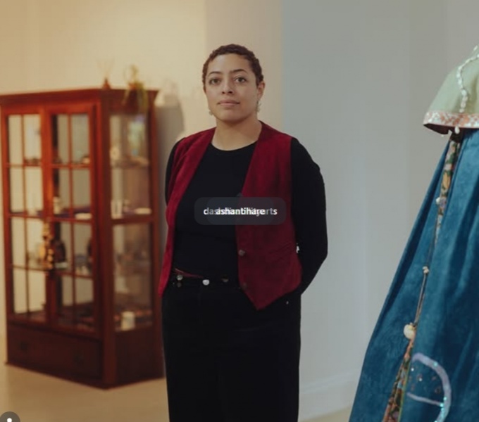 Curator Ashanti Hare stands in a white-walled gallery space. They are captured from the knees upwards. In the background to the left is a wooden glazed display cabinet, and in the foreground to the right is a blue, green and cream textile costume.