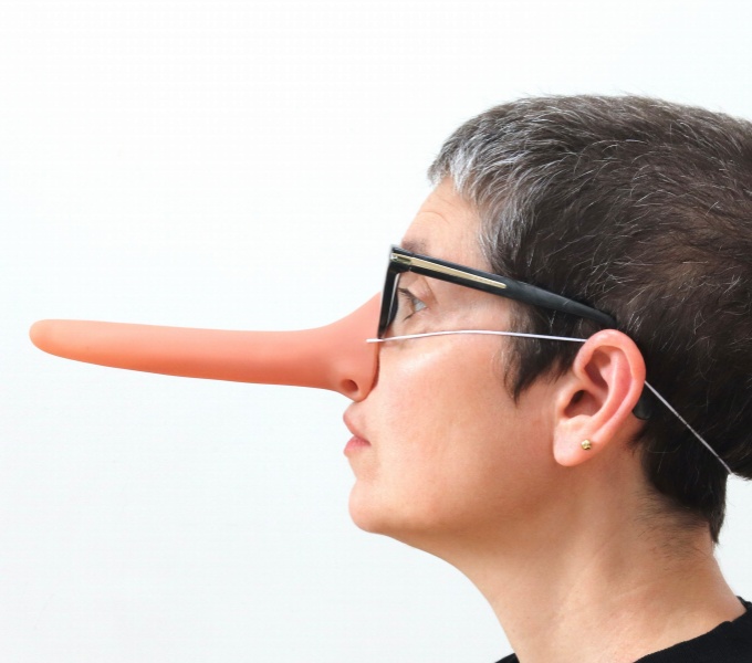 A side view of Sonia's head. She faces to the left. She has short dark grey hair and wears glasses. On her nose she wears a long costume pinocchio like nose. The background behind her is white. 