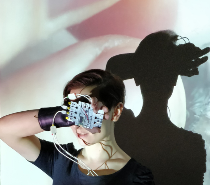 A photograph of the head and shoulders of a person brightly lit from the front, casting a dark shadow on the wall behind them. On the wall an enlarged close-up image of two fingers is projected. 