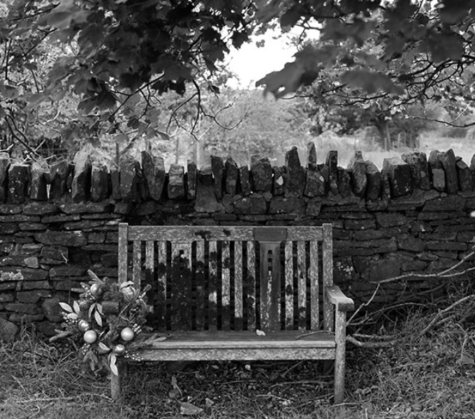 A black and white photograph of a wooden bench against a traditional dry stone wall.  Above is a leafy canopy of plane trees. The grass around the feet of the  bench is long and untamed and attached to the arm of the bench is a large floral arrangement.