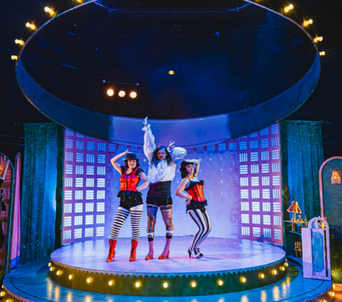  A photo of three performers in colourful costumes dancing on a circular, illuminated stage under a large ring of lights suspended from the ceiling. The theatrical set resembles a futuristic nightclub or cabaret venue with neon signage.