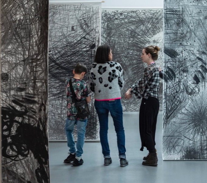 Image shows three people standing amongst black and white suspended drawings.