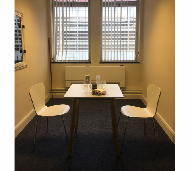 A photograph of the interior of a small room with large blinded windows at the rear, a window to the left and a table in the centre with a chair at either side of it. On the table there are glasses, a vase of water and biscuits on a plate.