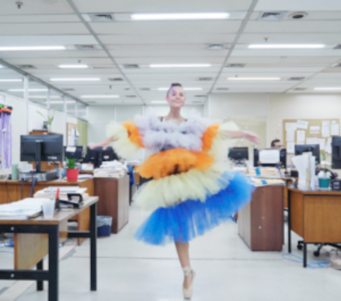 woman wearing a colourful multi-layered frilled dress and ballet shoes dances in an office space illuminated with strip lights