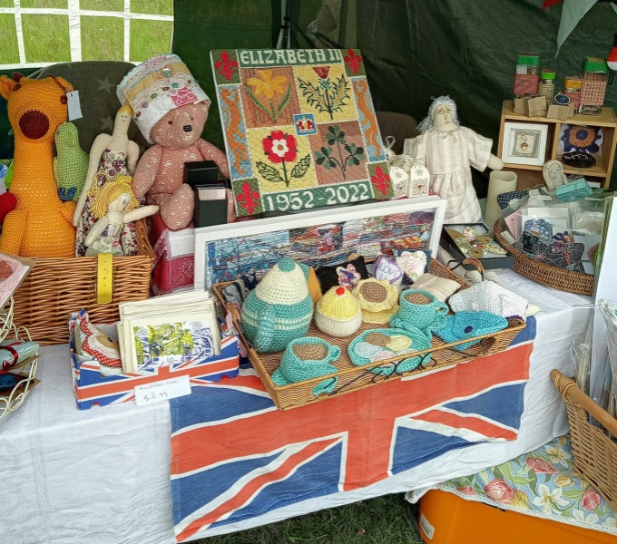A stall covered in a white table cloth and union jack flag. The table is full with hand crafted crotchet work. Including tea set, dolls and animals, as well as handmade card and other crafts.