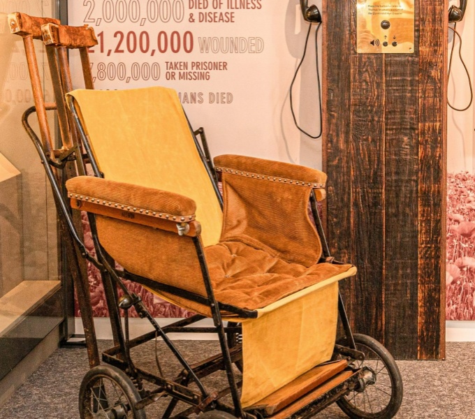 A cropped image of a vintage wheelchair. With painted black frame and yellow velvet seating cushions. The wheels are much smaller than todays design. Resting between a handle to the rear and the back rest are a pair of wooden crutches.