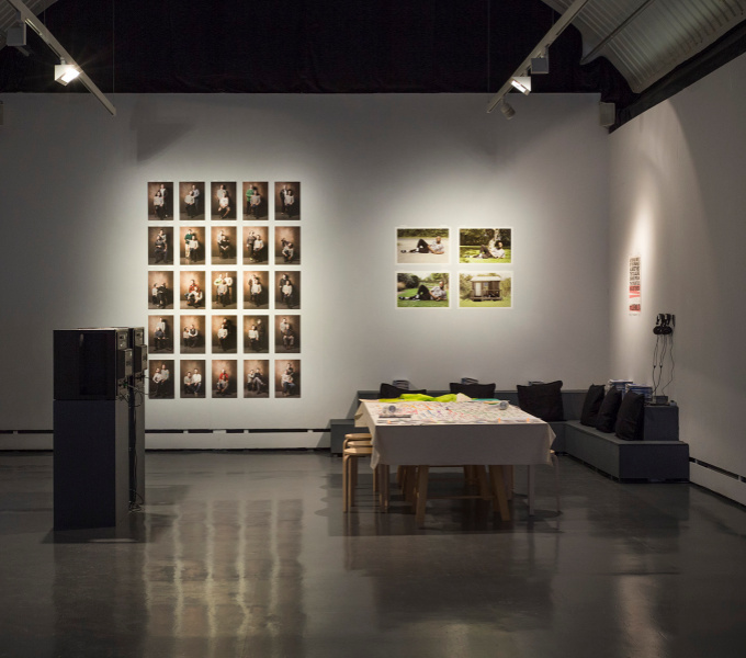 Exhibition space at Wysing Arts Centre. Photographic prints are on the far wall, while a corner sofa takes up the right hand far corner, headphones hang on the wall, a table with stools is in front of the sofa.