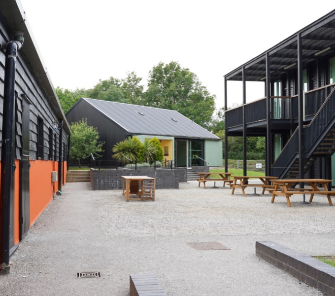 Courtyard space at Wysing Arts Centre. Picnic tables are in the central gravelled area, on the left is an orange and black building, behind is a grey and green building, on the right is a black metal 2 story building. 