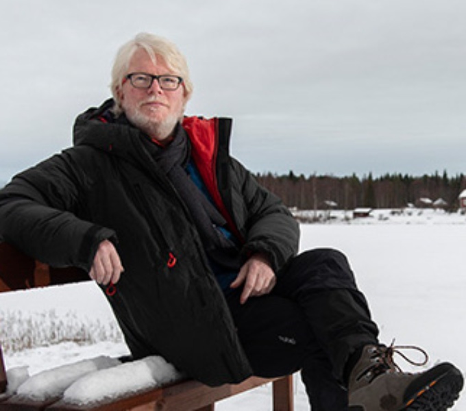 Aidan sits on a bench in a snow covered landscape. He wears a heavy dark coat, dark trousers and walking boots. 