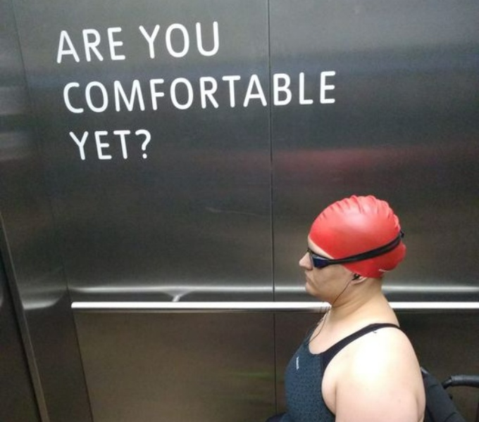 The steel interior of a lift. A woman wears a red swim cap, racing swimming costume and goggles is seated in a wheelchair. On the wall of the lift are the words: Are You Comfortable Yet?