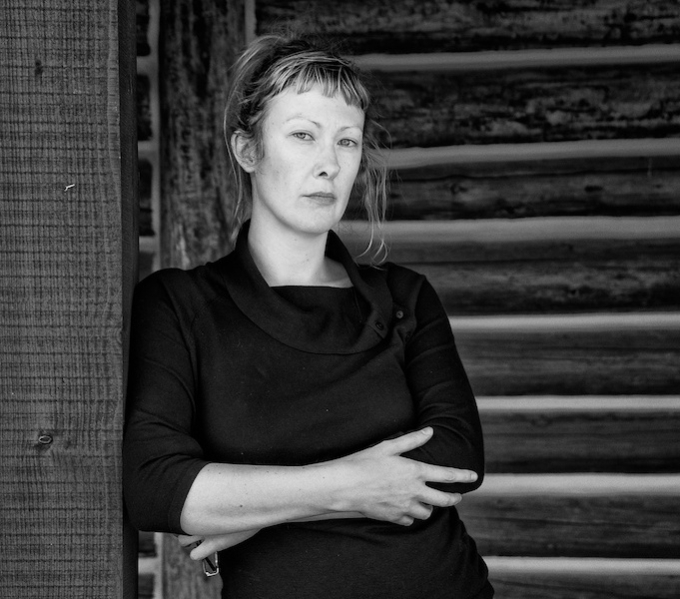 A black and white photographic portrait of Anna Berry, she is leaning against a wooden post and looking directly at the camera. Her arms a folded across her body.