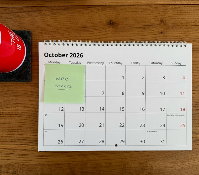 A view from above of a wooden desk with a monthly calendar opened on the October page.  At the top of the calendar, on a green post-it note reads NPO starts. To the left of the calendar is a red and white mug.