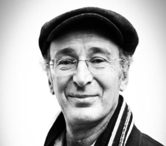 A black and white photographic portrait of Mike Layward; he wears glasses, a peaked hat and a striped scarf.
