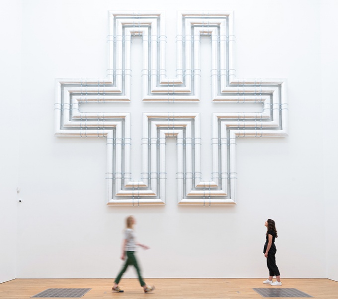 Two people stand in an art gallery, observing a large, abstract cross sculpture on a white wall.