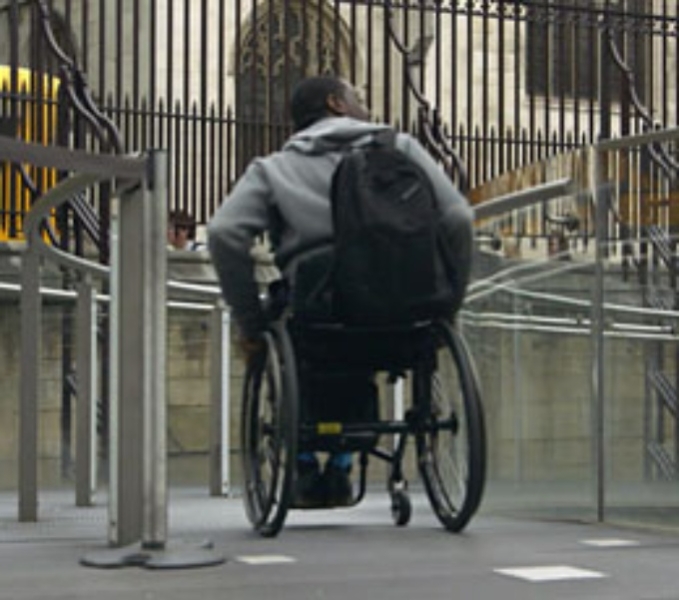 A wheelchair user navigates a city street. They wear a grey hooded sweatshirt and a black rucksack, and are approaching a ramp with glazed panels.