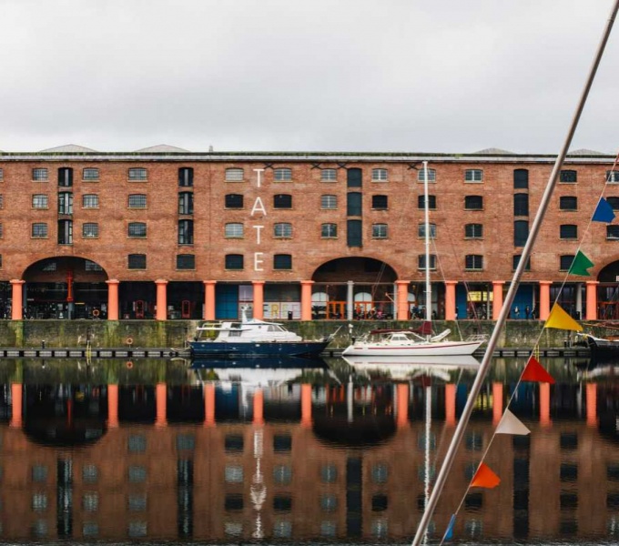 The Tate Gallery at Liverpool docks. Red brick building with painted red pillars below stands next to the waters edge. There are two small boats in the dock below.
