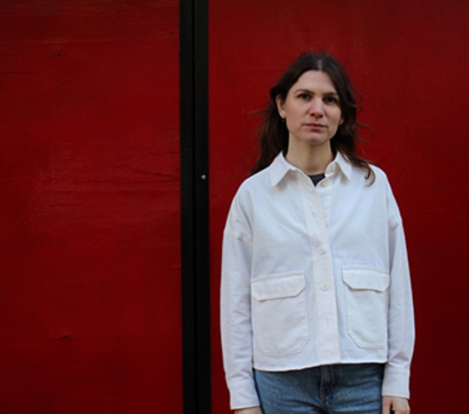 Hannah stands to the right of the photo in front of a deep red background. She wears a white shirt and blue jeans and has dark brown shoulder length hair. 