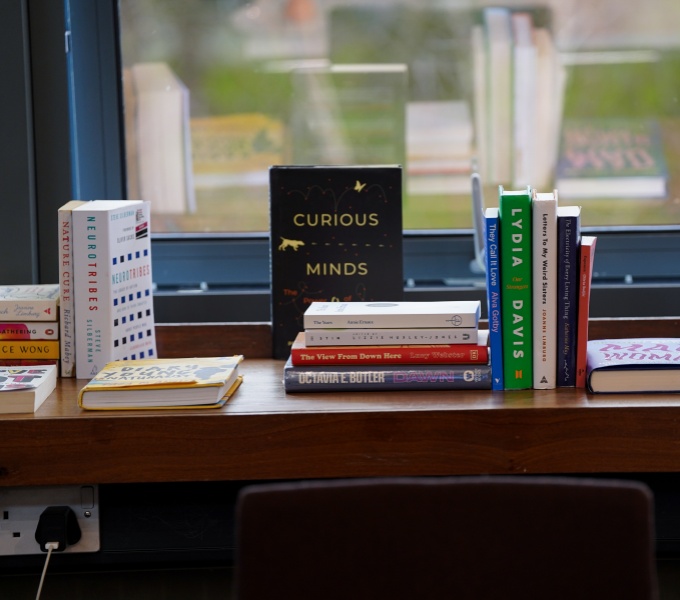 A number of books arranged on a ledge in front of a large window. Some books are stood on end whilst others are stacked sideways. Beyond, the view of lush green parkland and trees is out of focus and blurry.